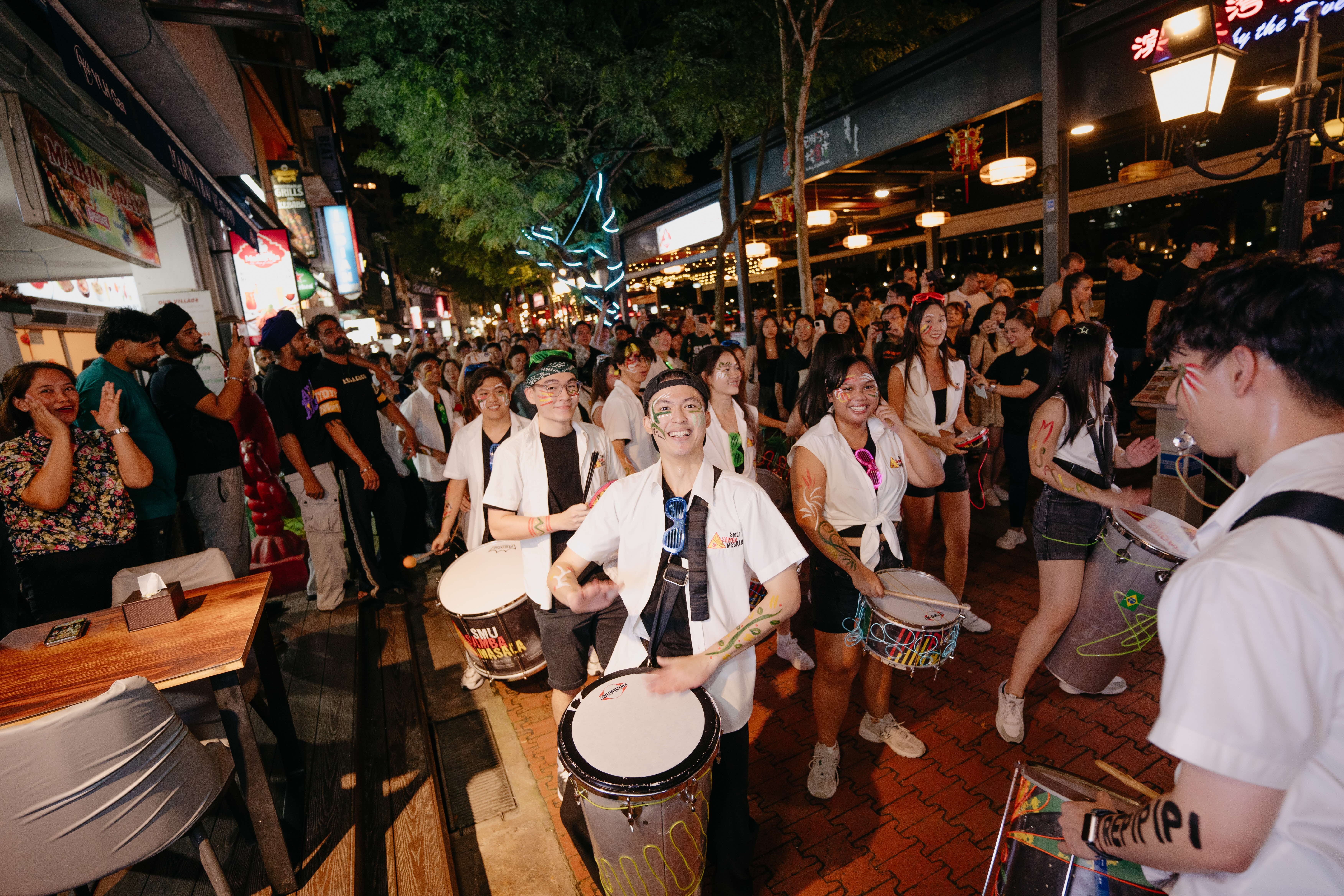 Night street scene of a parade with drummers and onlookers, people wearing face paint and bright lights.
